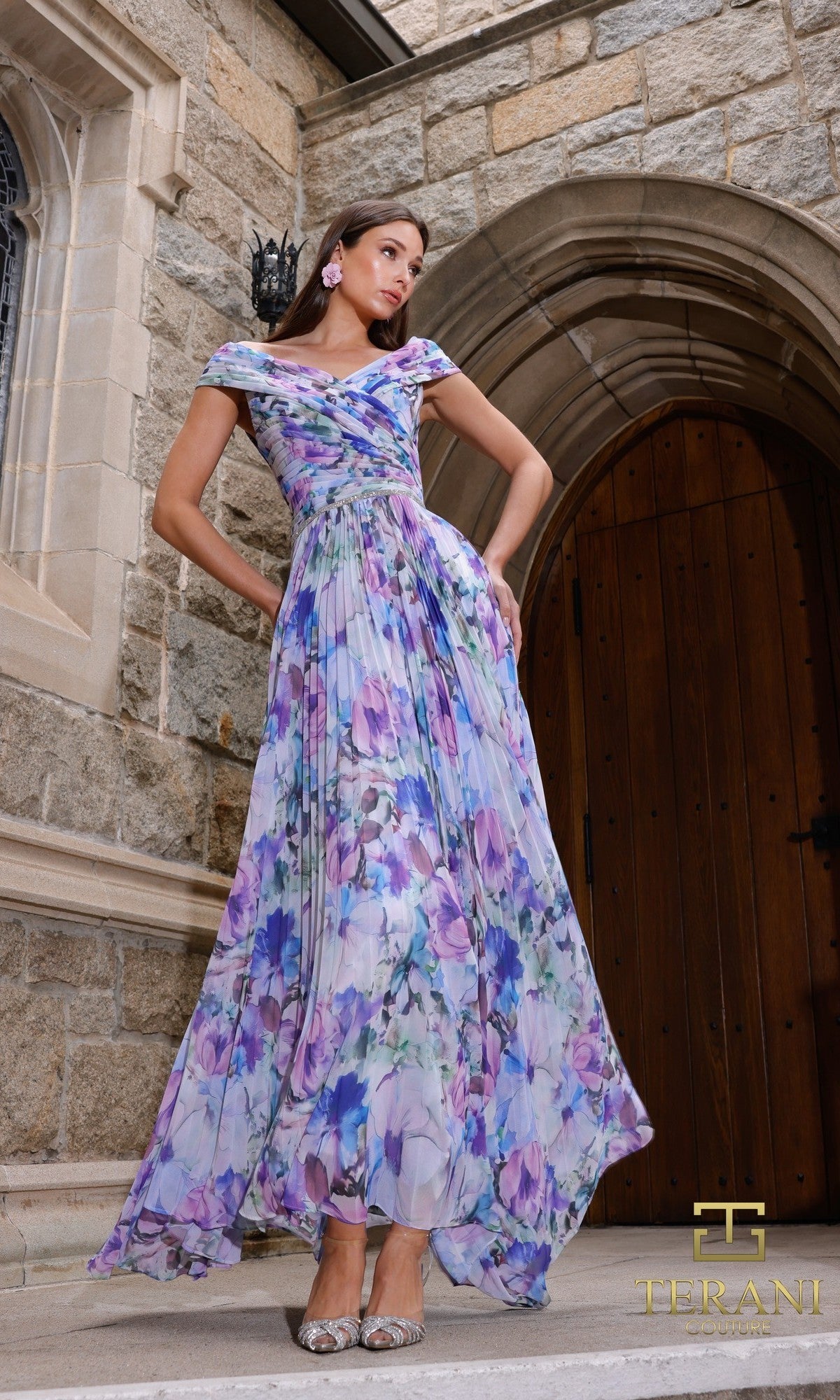 A woman stands on stone steps before an arched wooden door, wearing the Terani 261C6641 long formal dress in blue purple. She pairs it with silver shoes. The image displays the Terani logo in the bottom right corner.